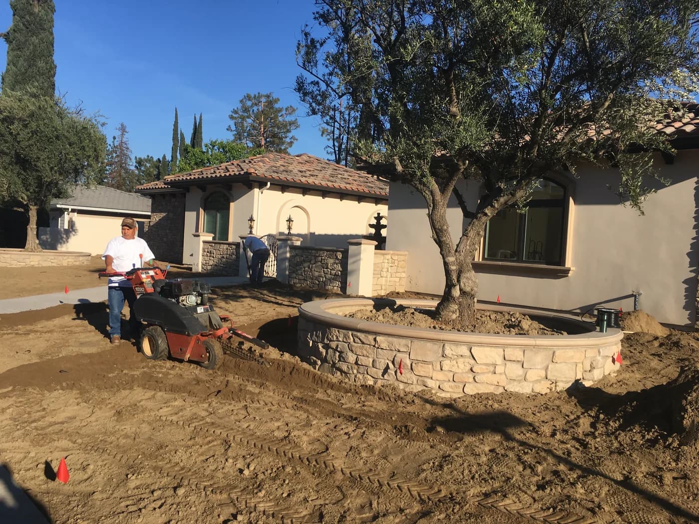 A crew member grading a new front yard with a walk-behind trencher beside a stone planter, the ground still bare dirt